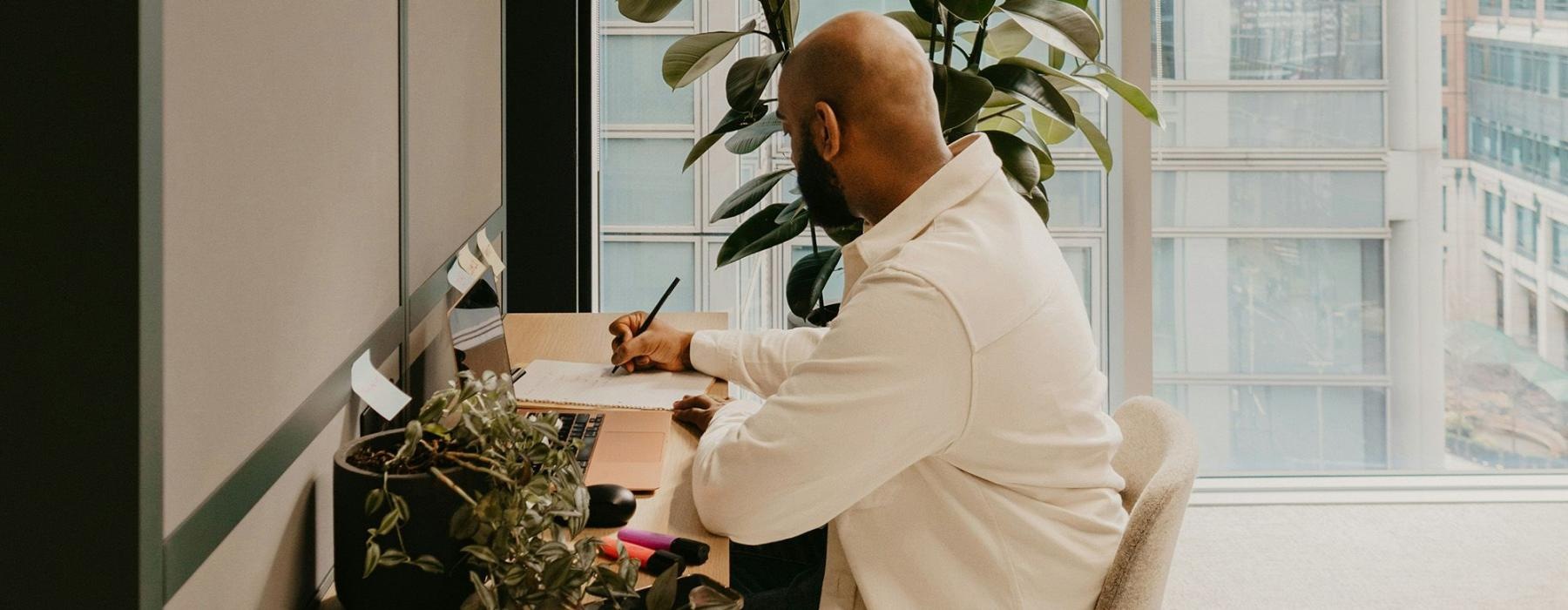 a man writing at a desk next to plants and a window