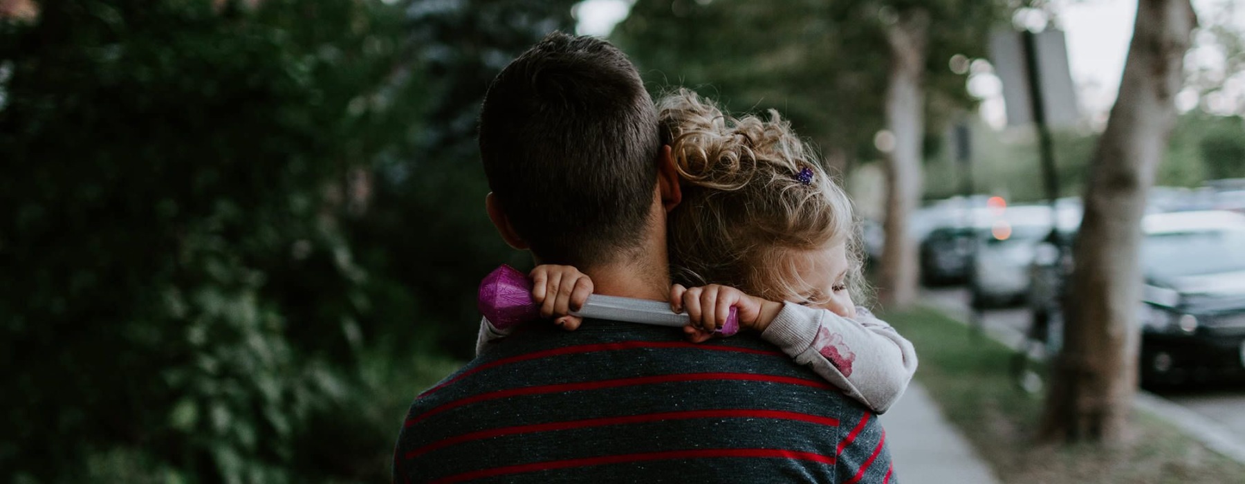 tired little girl rests her head on her father's shoulder as he carries her down the city sidewalk